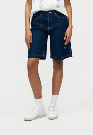 Persoon die donkerblauwe denim knielange shorts draagt, een wit mouwloos shirt met patroon, witte sokken en witte sneakers tegen een effen achtergrond.