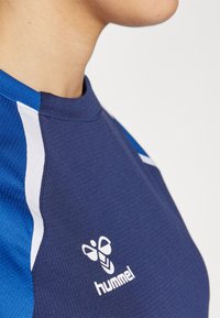 Lähivõte inimesest, kes kannab mereväesinist ja sinist Hummeli spordisärki valge logoga rinnal, ühevärvilise hele tausta ees.