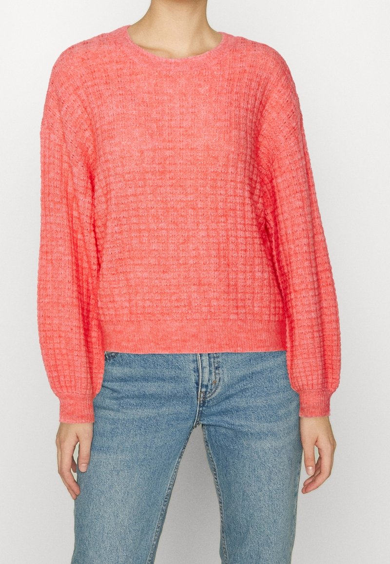 Koralle Strickpullover mit strukturiertem Muster, Rundhalsausschnitt und leicht puffierten langen Ärmeln. Kombiniert mit blauen Denimjeans.