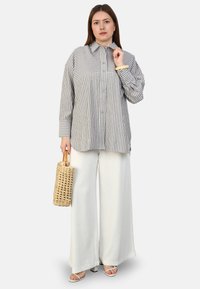 Chemise rayée à boutons grise et blanche avec des manches longues ; associée à un pantalon blanc ample. Les accessoires comprennent un sac tissé et des sandales.
