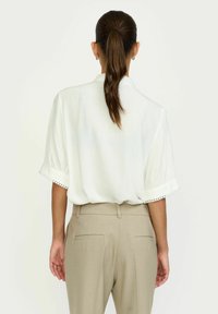 Bijela bluzica s kratkim, preklopljenim rukavima koja ima dekorativnu obrubu, u kombinaciji s bež krojenim hlačama. Glatka tekstura i opušteni kroj.