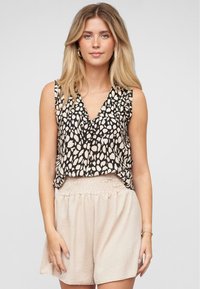 Femme aux longs cheveux blonds portant un haut sans manches à motifs noir et blanc et un short taille haute beige clair devant un fond uni.