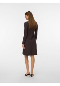 Smeđa haljina dugih rukava sa prilagođenim korzetom i raširenom suknjom, izrađena od mekog materijala. Hemline je do sredine leđa, a dopunjena je sandalama s otvorenom petom.