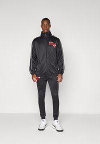Black satin tracksuit with red Chicago Bulls logo, featuring white side stripes, a zip-up jacket, and matching tapered pants. White athletic shoes.