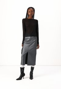 Haut transparent noir à manches longues avec un léger paillettes, associé à une jupe grise imitation satin arrivant au genou avec une fente à l'avant et des poches, complété par des bottes à talons noirs.