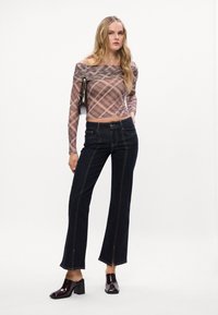 Dolga plaid majica v mauve odtenkih, visok ovratnik, v kombinaciji s temno modrimi širokimi kavbojkami z kontrastnim šivanjem in razcutimi robovi spredaj, obuta v sijajne mule.