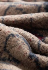 Coperta di lana con superficie morbida e testurizzata; base beige con motivi astratti blu e rossi; pieghe stratificate che creano profondità e volume.