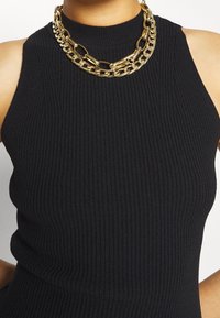 Top smanicato nero a coste con design a collo alto, caratterizzato da una collana a catena d'oro a più strati come accessorio. Texture morbida e forma aderente.
