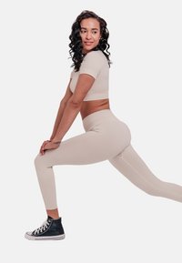 Beige crop top and leggings made of stretchy fabric, worn with black high-top sneakers. The model is performing a lunge stretch.