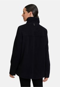 Donkerblauwe jas met hoge kraag, oversized pasvorm en minimalistisch ontwerp. Zachte textuur met een gecentreerde rugnaad en merklabel bij de hals.