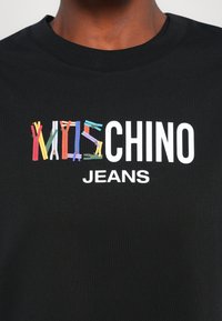 Juodas medvilnės marškinėliai su spalvingais užtrauktukų dizainais, įkomponuotais į tekstą "MOSCHINO JEANS" balta spalva.