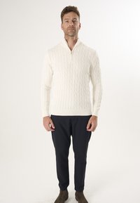 Cremfarbener Kabelstrickpullover mit hohem Kragen und halbseitigem Reißverschluss. Kombiniert mit dunkelblauen Hosen und braunen Stiefeletten.