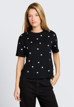 Junge Frau mit langen blonden Haaren, die ein schwarzes T-Shirt mit kleinen weißen Gänseblümchenmustern und schwarze Hosen trägt, steht vor einem schlichten Hintergrund.
