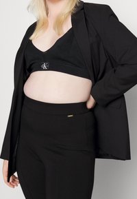 Musta bralette CK-logolla mustan bleiserin alla, paritettuna korkeavyötäröisten mustien housujen kanssa, joissa on pieni metallinen tunnus. Sileä pinta, istuva muotoilu.