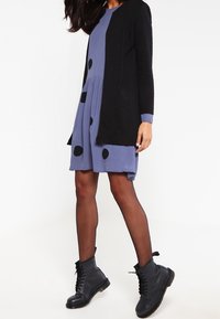 Mustanvärinen neulejakku sinisen mekon päällä, jossa on suuret mustat pilkut. Mekossa on levenevä hame ja pitkät hihat. Mustat nauhakenkäsaappaat.