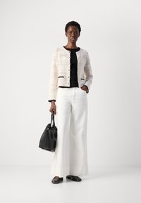 Cardigan bianco lavorato a maglia con bordi neri, indossato sopra un top nero. Pantaloni bianchi a gamba larga, borsa nera e scarpe nere completano l'outfit.