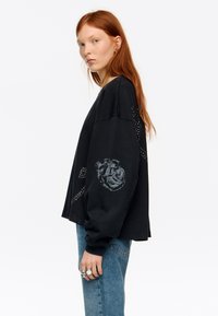 Sudadera negra de gran tamaño con un gráfico de rosa en la manga izquierda y detalles de tachuelas a lo largo del hombro, combinada con jeans azules.