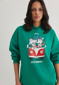 Green hoodie features a graphic of cats in a red car, with a “Join the Cute Parade” slogan. Soft, casual fabric, relaxed fit.