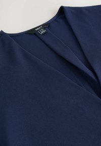 Blusa blu navy con design a V, realizzata in tessuto morbido. Presenta morbide pieghe all'escollatura e l'etichetta del marchio visibile all'interno.