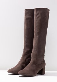 Bottes cuissardes en daim marron avec un bout pointu et un talon carré. Texture lisse, minimalisme des éléments métalliques et design épuré.