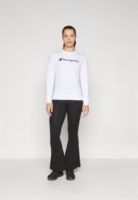 Sweatshirt blanc à col rond avec logo "Champion", associé à un pantalon noir évasé et des chaussures de sport noires. Design simple et épuré.