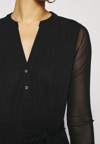 Frau in einem schwarzen Kleid mit durchsichtigen langen Ärmeln, einem runden Kragen, zwei kleinen Knöpfen und einem gebundenen Taillengürtel.