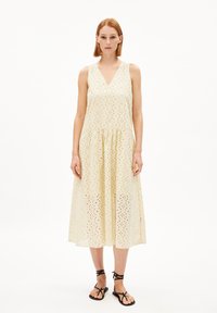 Helles gelbes ärmelloses Kleid mit V-Ausschnitt, ausgestattet mit Lochmuster und einem ausgestellten Rock. Kombiniert mit schwarzen Riemchensandalen.