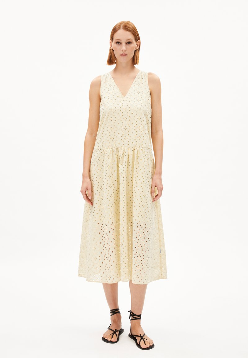 Helles gelbes ärmelloses Kleid mit V-Ausschnitt, ausgestattet mit Lochmuster und einem ausgestellten Rock. Kombiniert mit schwarzen Riemchensandalen.