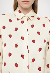 Cremfarbendes Hemd mit Knopfleiste, das ein Muster von roten Erdbeeren aufweist. Glattes Material mit klassischem Kragen und dezenter Logo-Stickerei am Saum.