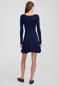 Marineblauwe geribbelde gebreide jurk met lange mouwen, ronde rug, uitlopende rok en een gladde textuur. Gestyled met eenvoudige bruine ballerina's, focus op de details van het kledingstuk.