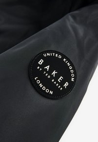 Černá látka s hladkou texturou obsahuje kulatý černý záplatu s bílým textem "BAKER by TED BAKER" a "UNITED KINGDOM LONDON".