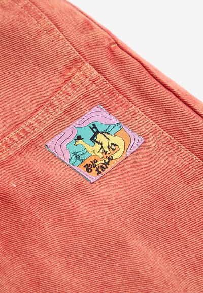 Tissu orange vif avec des coutures surpiquées et un petit patch coloré représentant un dinosaure dessin animé portant un chapeau dans une scène de désert.