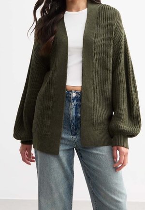 Femme portant un cardigan en maille épaisse vert foncé sur un crop-top blanc et un jean bleu clair taille haute, debout avec une posture détendue.