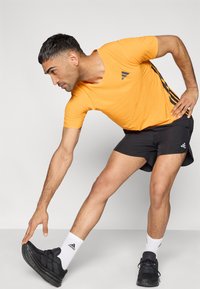 Πορτοκαλί αθλητικό μπλουζάκι με υφή κουκίδων, κοντά μανίκια, συνδυασμένο με μαύρο σόρτς. Το μοντέλο τεντώνεται προς ένα μαύρο αθλητικό παπούτσι, φορώντας λευκές κάλτσες.