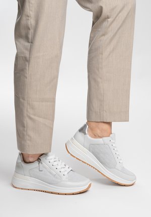 Personne portant un pantalon beige et des baskets blanches à semelle compensée avec fermeture éclair latérale et semelles en caoutchouc naturel, debout sur une surface blanche.