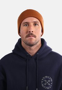 Un bonnet côtelé marron porté avec un hoodie bleu marine arborant un logo circulaire. Le bonnet a un ajustement serré avec une finition texturée.