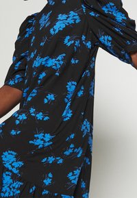 Černé šaty s krátkými nabíranými rukávy, zdobené modrou květinovou vzorovanou látkou s texturou. Rozšířený spodní lem dodává šatům lehkost a pohyb.