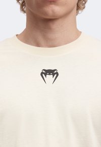 Νεαρό άτομο φοράει ένα κρεμ T-shirt με ένα μικρό μαύρο στυλιζαρισμένο λογότυπο κεφαλής φιδιού στο κέντρο κοντά στη λαιμόκοψη.