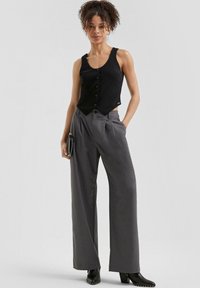 Zwarte mouwloze top met knoopdetails, gecombineerd met grijze broek met brede pijpen. Model houdt een zwarte clutch met gespikkelde accenten vast.
