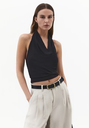 Donna con lunghi capelli castani indossa un top nero halter, pantaloni beige plissettati a vita alta, cintura nera con fibbia dorata, mani nelle tasche.
