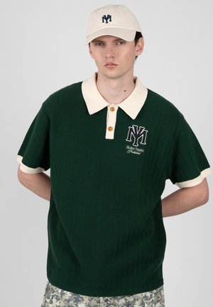 Mladý muž nosí tmavě zelené polo tričko s krémovým límcem a manžetami, krémovou čepici a maskáčové kalhoty, stojí s rukama za zády.