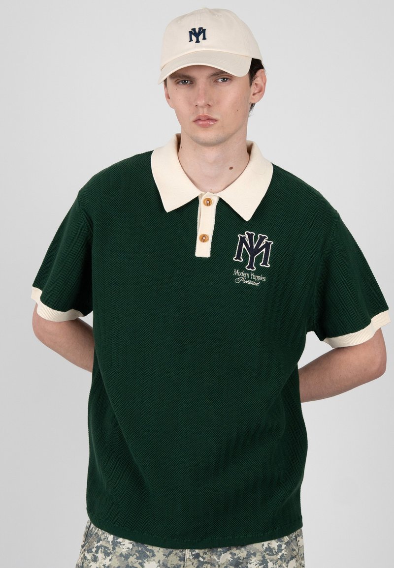 Νέος άνδρας φορώντας σκούρο πράσινο πόλο με εκρού γιακά και μανσέτες, εκρού καπέλο και παντελόνια παραλλαγής, στέκεται με τα χέρια πίσω από την πλάτη.