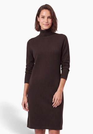 Une femme porte une robe en maille marron foncé, longueur genou, à col montant et manches trois-quarts, sur fond blanc.