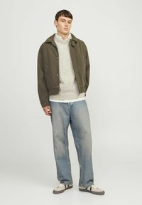 Oliivroheline vikinahkade villakate üle heleda halli sviitri, kombineeritud lõdva sinise teksapükste ja beežide tossudega, neutraalne taust.