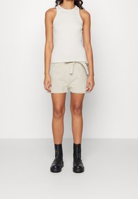 Top sin mangas de color claro combinado con pantalones cortos beige que tienen una cintura atada y bordes deshilachados. Botines negros completan el atuendo.