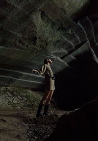 Una persona in un vestito marrone e stivali neri si trova su un terreno roccioso all'interno di una grotta buia, guardando verso l'alto verso un soffitto di pietra grezza.