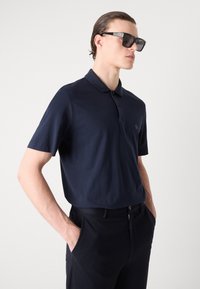 Jūros laivyno medvilnės polo marškiniai su trumpomis rankovėmis ir apykakle, su mažu logo. Pora su juodomis kelnėmis, fone – šviesiai pilkas.