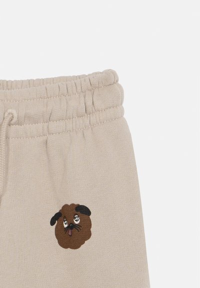 Beige sporta bikses no mīksta auduma, ar izšūtu karikatūras suņa seju kreisajā celiņā, melniem akcentiem un auklu jostu.