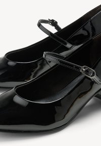Svarta lackskor med rund tå, låg klack och tunna justerbara remmar. Slät yta med en blank finish.