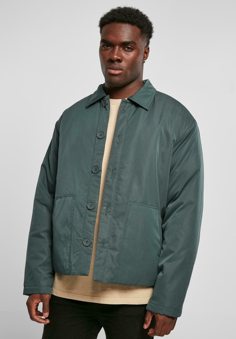 Mørkegrønn, middels tung jakke med knappestolpe, spiss krage og to frontlommer. Stoffet ser glatt ut med en matt finish.